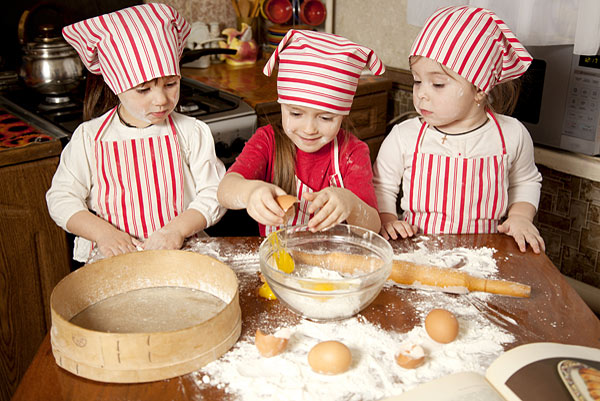 Drei Kinder in Kochmützen und Schürzen backen. Eines bricht ein Ei in eine Schüssel, während Mehl und Eier auf dem Tisch liegen.